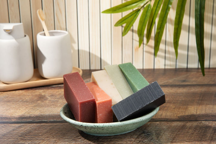 Six mini travel soap bars in a variety of colors, displayed in a green dish on a wooden surface. Ideal for shampooing color-treated hair, these paper soap shampoo and condition bars are perfect for travel. Ceramic containers and a toothbrush are in the background.